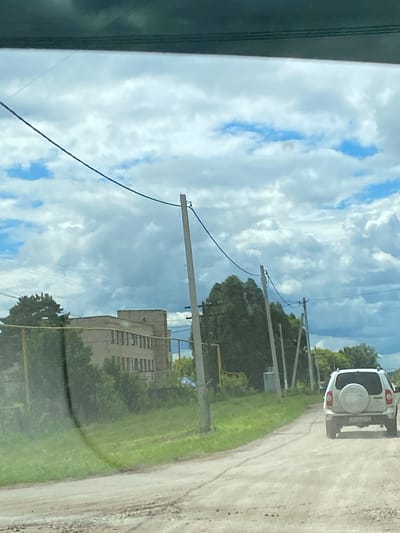 The image shows a white SUV driving on a dirt road in a rural area. The SUV is in the foreground, and the road is dusty. On the left side of the road, there is a building with a flat roof and several windows. There are also trees and power lines in the background. The sky is cloudy with patches of blue. The location appears to be outside of the city of Nadezhdino, Russia.