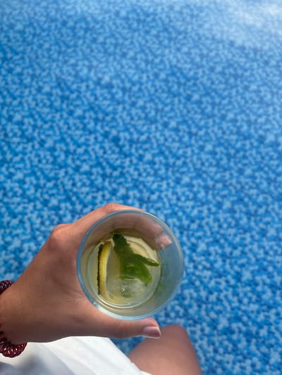 A hand is holding a clear glass with a drink, ice, a lemon slice, and mint leaves against a blue, speckled background that appears to be the bottom of a swimming pool. The glass is in the lower left quadrant of the image. A portion of a person's leg is visible in the lower right quadrant, wearing white clothing. The blue background is likely the tiled surface of a swimming pool, common in residential or public leisure facilities within the Tonbridge and Malling borough of the United Kingdom.
