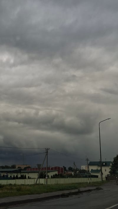 The image depicts an overcast sky over a street scene in Smalyavichy, Belarus. The sky is filled with heavy, grey clouds, suggesting impending rain or a storm. In the foreground, a wet asphalt road is visible, with patches of discolored pavement. Beyond the road, a strip of green grass and some vegetation lines a light green fence. Behind the fence, several buildings are visible, including structures with red roofs and a white building with a green roof. Electrical poles and wires are present throughout the scene, and a street lamp stands on the right side of the image. The overall impression is one of a somber, possibly gloomy day.