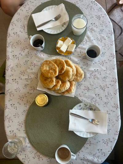 A top-down view captures a breakfast setting on a round table covered with a floral patterned tablecloth in Parvomay, Bulgaria. The table is set for at least two people, with two cups of coffee, a glass of milk, a small plate of cheese cubes, a plate of fried dough pastries, a jar of jam, and two place settings each with a plate, napkin, and fork. One place setting is partially visible at the top of the frame, with a fork resting on a napkin. Another place setting is at the bottom, with a fork on a napkin. A person's leg is visible at the top left corner of the image. The lighting suggests it is daytime. The environment appears to be an indoor dining area.