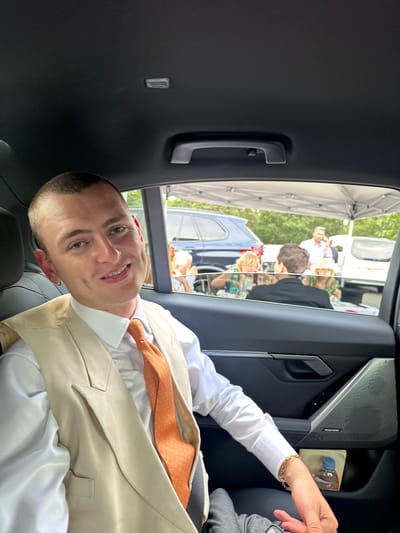 A man is sitting in the back seat of a vehicle, smiling at the camera. He is wearing a white dress shirt, a cream-colored vest, and an orange tie. His hair is short and shaved. He has a gold hoop earring in his left ear and a gold bracelet on his right wrist. In the background, through the car window, there are people gathered under a white tent. There are several cars visible, as well as trees and foliage. The scene appears to be outdoors, possibly at an event. The location is Ascot, United Kingdom.