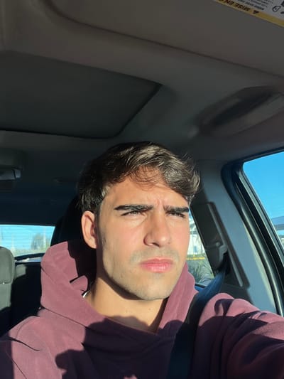 A close-up, selfie-style photograph shows a young man sitting inside a car. The car has an open sunroof, revealing a clear blue sky. The man has dark hair and is wearing a maroon-colored hooded sweatshirt. He is also wearing a seatbelt. The scene implies daytime and the location is inside a vehicle, and there is no evidence of Sarandí, Argentina.