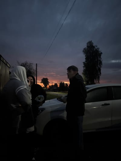 This is a medium shot, outdoor, eye-level, slightly angled photograph captured during twilight in the village of Ryabinino, Russia. The scene depicts four individuals gathered near a white car parked on a gravel surface. The sky is a dramatic gradient of deep blue, purple, and orange, indicative of sunset or sunrise.
In the foreground, on the left, a person wearing a light-colored hooded jacket with dark stripes on the sleeves is partially visible, facing away from the camera. To their right, a second individual in a dark hooded sweatshirt is also facing away, with their left hand raised to their face, appearing to be looking at something. Their right hand is holding a white object, possibly a cloth or a small bundle.
To the right of the first two individuals, a third person, a young male, stands facing left, wearing a black hooded sweatshirt and blue jeans. He is looking towards the other individuals, with his hands by his side. He is positioned in front of the white car.
Behind the group, a tall, dark tree stands silhouetted against the colorful sky. The car appears to be a sedan, with its driver's side door visible and slightly ajar. A building with corrugated metal siding is visible to the far left of the frame. In the background, across a dirt road, are more trees and what appears to be houses, suggesting a rural or village setting. Power lines are visible stretching across the upper portion of the sky. The overall mood is casual and communal.