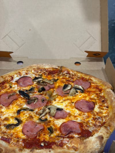 An overhead, slightly angled close-up shot shows a pizza in an open pizza box. The pizza has a golden-brown crust and is topped with melted cheese, red sauce, sliced red sausage, and sliced brown mushrooms. The pizza box is made of light brown cardboard. The background is blurred and appears to be a dark, flat surface. No people or identifiable landmarks are present in the image, and therefore, the location of Korolyov, Russia cannot be determined from the visual information provided.