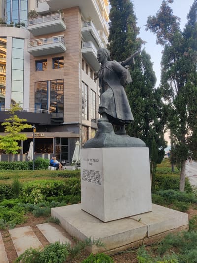 The image shows a bronze statue of a woman holding a rifle, standing on a stone pedestal in an outdoor urban setting. The statue is positioned in front of a modern building with glass facades and balconies, and surrounded by greenery, including cypress trees and manicured bushes. The pedestal has an inscription in Greek, which reads "ΓΥΝΑΙΚΕΣ ΤΗΣ ΠΙΝΔΟΥ 1940" and a quote attributed to the women of Pindos. In the background, people are seen sitting at tables outside a cafe. The overall scene suggests a public monument or memorial within the city of Thessaloniki, likely in the Municipal Unit of Thessaloniki, Greece. The statue is a historical monument, likely commemorating women's participation in war or resistance, given the inscription and the rifle. The environment is well-maintained, with a mix of natural elements and urban architecture.