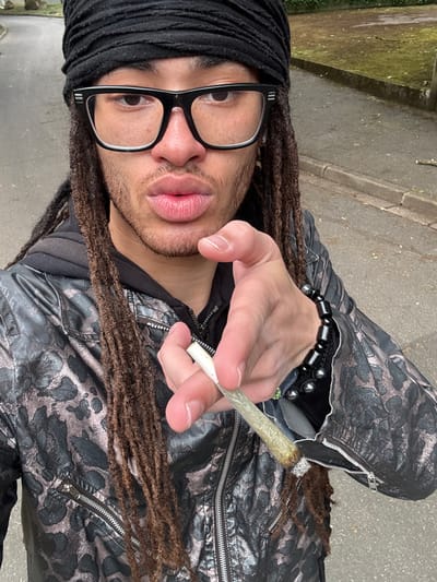 A man with dreadlocks, wearing a black head wrap and glasses, is holding a lit joint in his right hand. He's wearing a dark patterned jacket with zipper details. The background appears to be an outdoor urban environment, possibly a sidewalk or street in Les Ulis, France. The exact location cannot be determined from the image.