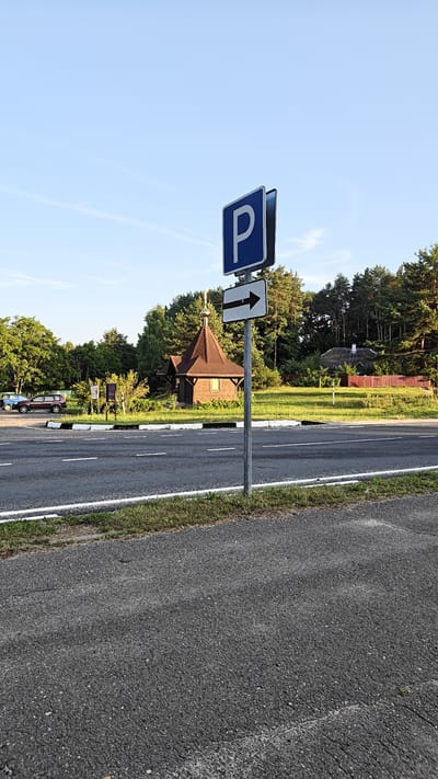 The image captures a view of a road and its surroundings on a sunny day in the village of Нарацкі сельскі Савет, Belarus. A blue and white parking sign with a white arrow pointing to the right is prominently displayed on a metal pole in the foreground. The road, paved with asphalt and bordered by white lines and a strip of grass, runs horizontally across the lower portion of the frame.
In the middle ground, behind the road, a small, traditional wooden building with a dark brown, steep, conical roof and a golden cross on top is visible. Adjacent to this building, a few cars are parked on a grassy area. Further in the background, a dense line of pine trees forms a backdrop, and a red wooden fence can be seen. The sky above is a clear, light blue with a few wispy clouds. The scene depicts a typical rural or semi-rural setting, likely a point of interest or amenity for visitors to the area.