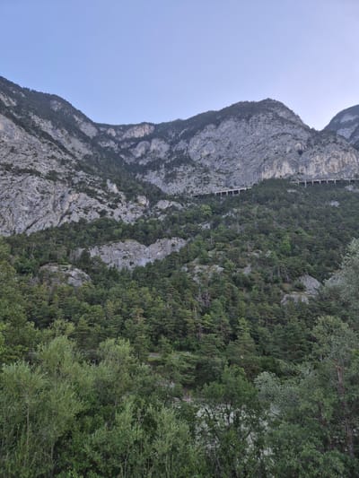 The image depicts a mountainous landscape with a clear, pale blue sky. The mountains are steep and rocky, with patches of dense green forest covering their lower slopes. A man-made structure, possibly a road or railway viaduct with arched supports, is visible traversing a section of the mountainside higher up. The foreground is filled with lush green foliage, including trees and bushes, suggesting a natural environment. The scene appears to be a scenic view of a mountainous region, likely a natural park or a less urbanized area. The crops show closer views of the mountain terrain, forests, and the sky, highlighting the textures of the rocks and the density of the trees. There is no indication of human presence other than the viaduct and no specific actions or interactions are captured. The location is identified as Kematen in Tirol, Austria.