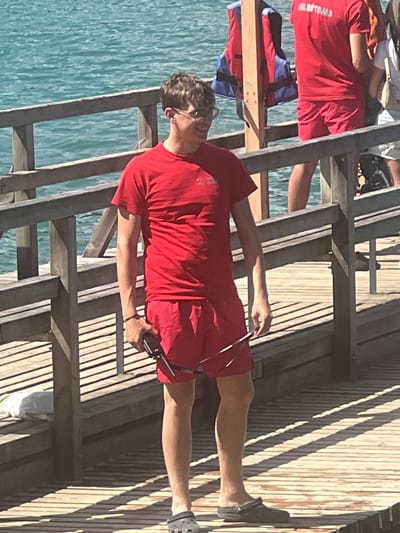 A group of people, possibly lifeguards, are on a wooden structure by a body of water. One person in the foreground, wearing a red t-shirt and red shorts, holds a device that appears to be a radio or walkie-talkie. This person is also wearing sunglasses and has on dark-colored clogs. Another person in the background is also wearing a red shirt. The wooden structure appears to be a pier or boardwalk with railings and steps. The water is a clear blue-green color, suggesting it is a sunny day. The sun is casting shadows across the wooden planks, indicating it is likely daytime. There is text visible on the red shirt of the person in the foreground, but it is difficult to read due to the angle and resolution. The overall scene conveys a sense of activity and preparedness, possibly related to water safety or recreation. The presence of the wooden structure and the clear water suggests an outdoor environment, possibly a lake, river, or coastal area.