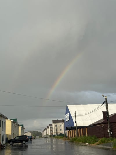 A double rainbow arcs across a cloudy, gray sky over a street in Murom, Russia, after a rain shower. The street is wet and reflective. On the left, several two-story residential buildings with yellow and white facades and green roofs line the street. A black SUV is parked on the left side of the street, and another blue car is visible further down. On the right side of the street, a large building with a white, tent-like roof and a blue section with Cyrillic text that reads "КРЫЛЬЯ" (Wings) is visible. Adjacent to it is a brown wooden fence and a street light on a wooden pole with numerous electrical wires. The overall impression is of a quiet, post-rain suburban or commercial area.