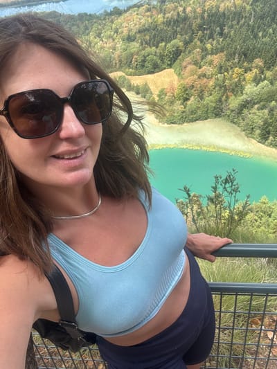 In this selfie, a woman with brown hair and sunglasses stands on a viewing platform, leaning on a dark green metal railing. She is wearing a light blue sports bra and dark blue shorts. Behind her, a vibrant turquoise lake is visible, surrounded by lush green trees and hills. The scene appears to be a scenic overlook, possibly at Lac d'Ilay, a natural lake in the Jura region of France, near the commune of La Chaux-du-Dombief. The lighting suggests it is daytime.
