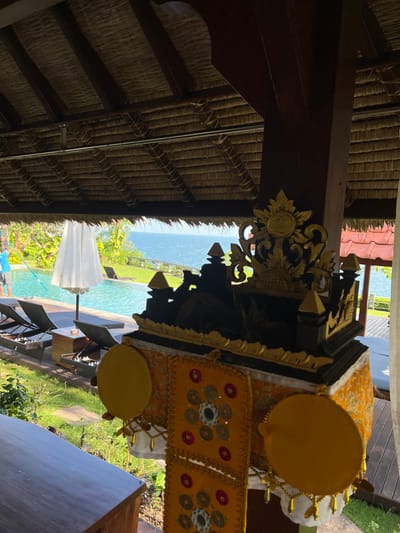 The image depicts an outdoor tropical resort scene in Uluwatu, Indonesia, viewed from beneath a shaded structure with a thatched roof. In the immediate foreground on the left, a dark wooden surface, possibly a table or counter, is partially visible. Centrally, a Balinese ceremonial object, likely a shrine or offering stand, is prominent. This object is dark-colored with intricate gold carvings, featuring stylized pagoda-like structures and decorative flourishes, including a sun-like emblem at its peak. It is draped in patterned yellow and orange fabric, adorned with multiple circular yellow discs and smaller, coin-like reflective embellishments arranged in geometric patterns. The overhead view reveals a densely woven thatched roof supported by dark wooden beams.
In the midground, a light blue infinity pool is visible, surrounded by a dark wooden deck. Several dark-colored lounge chairs with white cushions are arranged on the deck. A large, open white umbrella provides shade near the pool. To the left, a single individual wearing a light blue shirt is partially visible, actively engaged in cleaning the pool using a long pole.
The background features an expansive view of the blue ocean extending to the horizon under natural light. A manicured green lawn separates the pool deck from the ocean, bordered by a white railing. Distant lush green trees and foliage line the coastline, and a structure with red-tiled roofs is partially observable on the far right.