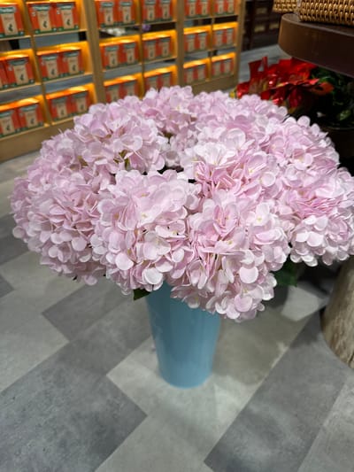 A close-up, eye-level shot focuses on a large bouquet of artificial pink hydrangeas, displayed in a light blue vase. The flowers are densely packed, with delicate, pale pink petals. The vase is positioned on a grey and white tiled floor, arranged in a diagonal plank pattern. In the background, out of focus, shelves are visible with rows of orange boxes, suggesting a retail environment. To the right of the flowers, a glimpse of a woven basket and a blurred red object, possibly part of a decorative display, can be seen. The overall setting appears to be indoors, likely in a shop or home decor store. There are no people or animals present in the image. The scene is static and well-lit, with the focus clearly on the floral arrangement. The specific location of Phu Thuy, Vietnam is not discernible from the visual elements in the image.