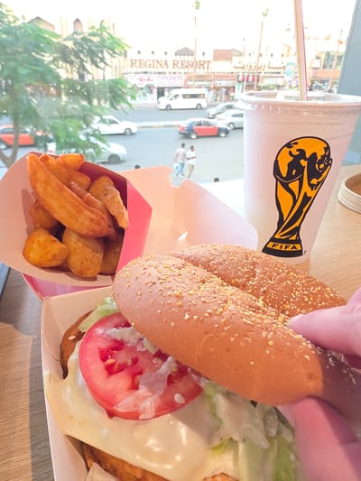 The image depicts a close-up view of a meal on a table, with a blurred background showing a street scene in Al Hadaba, Egypt. In the foreground, a burger is partially obscured by a hand, revealing lettuce, tomato, and cheese. To the left, a box of potato wedges is visible. A white cup with a FIFA logo and a straw stands to the right of the burger. The background shows a street with cars, buildings, and trees. A sign for "REGINA RESORT" is prominent on one of the buildings. The overall lighting suggests it is daytime.