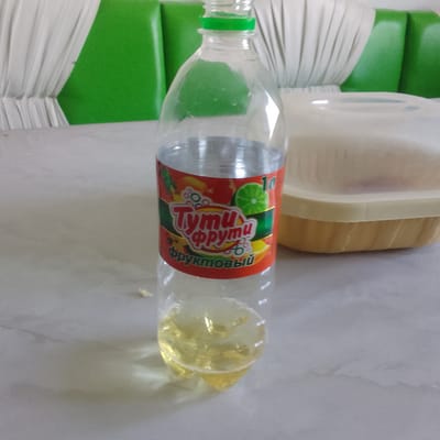 A clear plastic bottle filled with a yellow liquid stands on a light-colored, marbled surface. The bottle has a green cap and a label with red, green, yellow, and orange designs. The label features the text "Тути Фрути" (Tuti Fruti) and "фруктовый" (fruit) in stylized lettering, along with images of fruit slices and bubbles. To the right of the bottle is a light beige plastic container with a transparent lid, also on the same surface. In the background, green padded seating with white pleated fabric elements is visible. The overall scene appears to be indoors, possibly in a cafe or dining area. No people are present in the image. The location in Almaluu, Kyrgyzstan, is not discernible from the visual information provided.