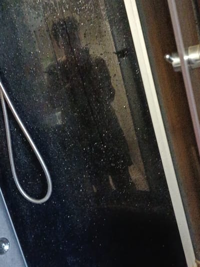 The image shows a reflection in a dark, speckled surface that appears to be a shower door with water droplets on it. The reflection is of a person, likely a woman, with dark hair, wearing dark clothing. She is holding a phone in her right hand and appears to be taking a selfie. The background of the reflection is blurred, but it suggests an indoor setting, possibly a bathroom. To the right of the shower door, a portion of a wooden surface and a chrome shower fixture are visible. The overall impression is a candid moment captured in a personal space. The lighting suggests it could be daytime, with light entering from an unknown source, illuminating the water droplets and creating specular highlights. There is no visible text in the image. The scene is intimate and personal, capturing a moment of self-documentation in what appears to be a private setting.