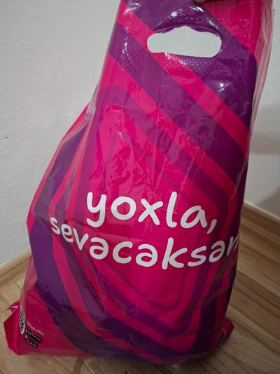 A bright pink and purple striped plastic shopping bag is leaning against a white wall and resting on a wooden floor. The bag has a cutout handle at the top. White text on the bag reads "yoxla, sevacaksan". At the bottom left of the bag, there are icons for the App Store and Google Play, and text in a non-English language. The scene appears to be indoors, likely a home or retail environment. No people are visible.