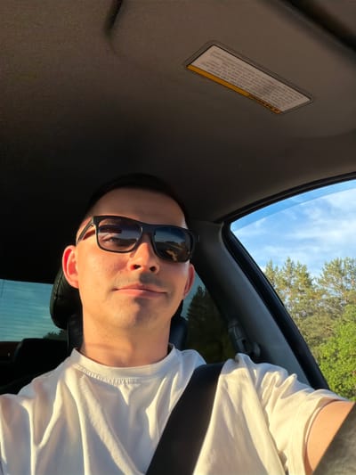 The image captures a person, likely male, from a low angle within a car's interior. He wears black sunglasses and a white t-shirt, looking directly at the camera with a neutral expression. The scene is illuminated by warm, golden sunlight hitting the right side of his face and shirt, suggesting late afternoon or early evening.
Above him, an open sunroof reveals a clear blue sky with sparse clouds and the tops of green trees, indicating an outdoor setting. The car's interior features a dark headliner. A visible warning sticker on the sun visor reads: "A WARNING. DEATH OR SERIOUS INJURY can occur. NEVER put a rearward-facing child seat in the front. The BACK SEAT is the safest place for children. ALWAYS use seat belts and child restraints. See the Car's Manual for further information and safety." The warning emphasizes child safety and proper use of car seats and restraints.
The person appears relaxed while in the vehicle, which is situated outdoors in Yus'ki, Russia, under sunny weather conditions.