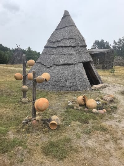 The image depicts an outdoor scene at an archeological reconstruction site in Камянюцкі сельскі Савет, Belarus. In the foreground, a vertical wooden post displays several clay pots of various sizes, some with intricate patterns. These pots are scattered around the base of the post and on the ground. Behind this display, a large conical hut with a thatched roof is prominent. To the right of the conical hut, a rectangular wooden structure with a thatched roof is visible, and further in the background, another conical structure and dense trees line the horizon. The ground is covered with dry grass and patches of soil. The sky is overcast.