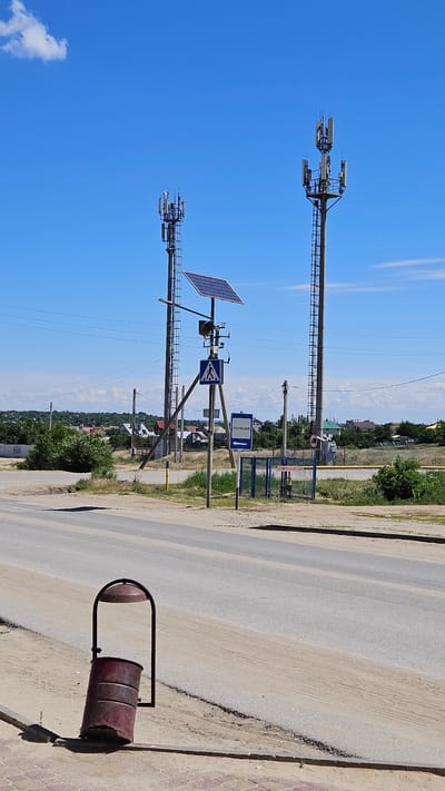 The image shows a street scene in Yerzovka, Russia. Two telecommunication towers are visible against a clear blue sky. The towers are equipped with antennas and solar panels. One of the towers also has a pedestrian crossing sign and a police station directional sign. A metal trash bin lies on the edge of the road. The road appears to be paved and runs parallel to the line of sight. Vegetation and buildings can be seen in the background.