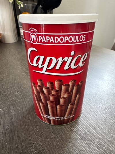 A tall, cylindrical red tin of Papadopoulos Caprice chocolate-filled wafer rolls sits on a grey wooden table. The tin has a white lid and a prominent "Caprice" logo in white cursive font. Below the logo, a cluster of chocolate wafer rolls is depicted. Text on the lower part of the tin reads "DELICIOUS WAFER ROLLS WITH HAZELNUT AND COCOA CREAM". The scene is indoors, with the table surface filling the foreground. No people or specific city locations are visible in the image.