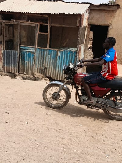 An adult male is observed seated on a red Bajaj Boxer motorcycle on an unpaved dirt surface in Zango, Nigeria. He wears a red, white, and blue short-sleeved shirt, dark shorts, and flip-flops, grasping the motorcycle handlebars and facing towards the right side of the frame. The motorcycle has a silver front fender and spoked wheels. In the background, a weathered building with a white corrugated metal roof and a facade constructed from blue corrugated metal sheets and wooden frames with mesh inserts is visible. A wooden door is present on the left side of this structure. An inscription is discernible above the blue panels. To the right, another structure made of lighter-colored material features a dark, open doorway. The ground consists of dry, light-brown earth, showing minimal scattered debris. The scene is under natural daylight, casting shadows.