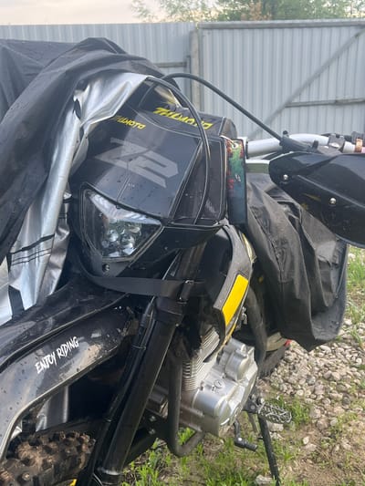 In Vasilyevo, Russia, a black motorcycle is partially covered by a black and silver tarp. The motorcycle has a black front fender with a yellow stripe and a black headlight assembly. A sticker on the side of the motorcycle reads "ENJOY RIDING" in white lettering. The motorcycle is parked on a gravel and grass surface, with a corrugated metal fence in the background. The sky is overcast.