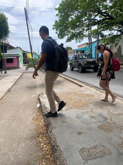 The image captures a street scene in Tacarigua, Venezuela. Two people are the main subjects. In the foreground, a person wearing a dark blue patterned short-sleeved shirt, tan pants, and black sneakers walks on a sidewalk, carrying a black backpack. Their head is tilted down, and their right arm is slightly extended. Behind them, a woman with long dark hair, wearing a floral print dress and light-colored sandals, walks on the same sidewalk. She has pink headphones on her ears and a bright pink backpack slung over her left shoulder.
The setting appears to be an urban or semi-urban environment. A black Jeep is parked on the street to the left of the woman. A green street sign with white text is visible near the Jeep. Colorful buildings, painted in shades of blue, yellow, pink, and green, line the street. Overhead, a network of power lines crisscrosses the sky, with a wooden utility pole prominent in the frame. Lush green trees, with their branches extending over the sidewalk and street, add to the scenery. The ground is a mix of concrete sidewalk and asphalt road, with scattered leaves and debris.
The lighting suggests it is daytime, possibly overcast or with diffused sunlight, as there are no harsh shadows. The overall mood appears casual, with the individuals seemingly going about their day. The visible text on the street sign reads "Sta. Ana" and "Juana Greco".