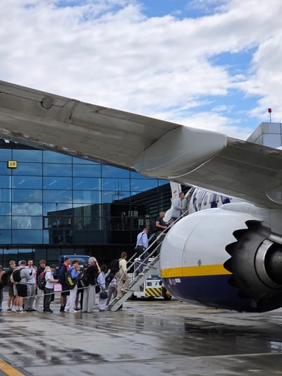 The image depicts a boarding scene at Balice John Paul II International Airport in Balice, Poland. A group of passengers, a mix of young adults and some older individuals, are lined up on a mobile airstair, preparing to board a Ryanair aircraft. The aircraft's engine is prominently visible on the right side of the frame, with its characteristic blue and yellow livery. Behind the aircraft and passengers is the glass facade of the airport terminal, reflecting the sky and clouds. The ground appears wet, suggesting recent rain. One passenger at the front of the line is ascending the stairs, while others wait their turn. The sky is partly cloudy with patches of blue. The overall scene captures the activity of air travel during what appears to be daytime.