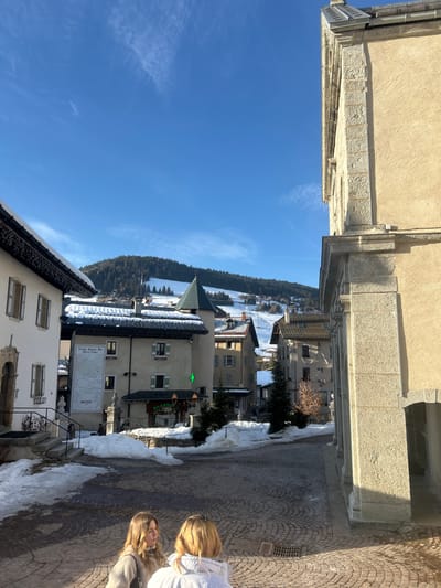 A picturesque village scene unfolds on a sunny day. Buildings are covered in snow, nestled against a backdrop of a forest-lined lake and clear blue sky. A cobblestone street lined with snow drifts leads to a building with a sundial on its wall. Two people are visible in the foreground, likely tourists. The perspective is from an elevated position, possibly a building's terrace.