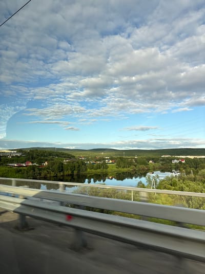 A scenic view of the city of Zverosovkhoz, Russia, is captured from a moving vehicle. The foreground features a blurred metal guardrail with a red mark on its lower section, suggesting motion. Beyond the guardrail, a calm body of water reflects the sky and the surrounding lush green landscape. The midground displays a residential area with houses nestled among trees, and a riverbank. In the background, rolling hills covered in dense vegetation stretch towards the horizon, under a sky filled with scattered white clouds against a bright blue expanse. Power lines are visible stretching across the sky. The overall scene depicts a peaceful, natural environment with signs of human habitation.