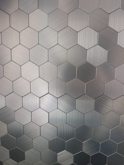 The image shows a close-up, eye-level view of a wall covered in brushed metal hexagonal tiles. The tiles are arranged in a honeycomb pattern, and the brushed texture creates a metallic sheen with visible lines of light reflection. The color palette is predominantly gray and silver, with subtle variations in tone across the tiles due to the lighting and the brushed finish. There are no people or other discernible objects or actions in the image. The background is entirely composed of these tiles. The image does not contain any information that would allow for its location to be identified as Naro-Fominsk, Russia.