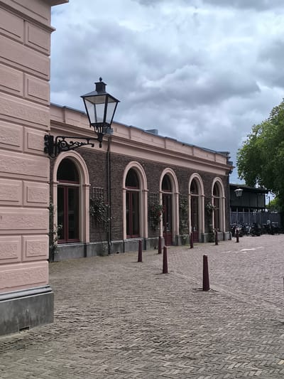 The image captures a street scene in Utrecht, Netherlands, characterized by distinctive architectural elements. A two-story building, brick on the ground floor and covered with a blush-colored brick wall, featuring a row of arched windows is prominently displayed. Decorative wrought-iron lampposts are mounted on the wall and stand on the sidewalk. In the distance, a bicycle rack can be seen holding multiple bicycles. The ground is covered with herringbone brick paving. A line of red traffic bollards parallels the facade. The sky is overcast.