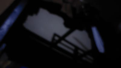 The image is a blurred, dark photograph that appears to be taken indoors. A diagonal blue-lit object is visible on the left side. A large, dark, irregular shape dominates the majority of the frame, with some lighter gray areas suggesting a surface or wall behind it. The overall impression is one of low light conditions and a lack of clear detail. The image does not provide any visual cues that would allow for the identification of its location in New Nyanya, Nigeria, nor does it depict any discernible people, subjects, objects, actions, or interactions.