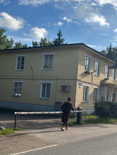 In Annino, Russia, a person wearing a brown robe and head covering walks past a yellow brick apartment building. The building has several windows with white frames, and a street sign reading "Sadovaya ulitsa 10" is visible on the exterior wall. A white and red striped barrier extends across the foreground. The sky is blue with scattered white clouds, and trees are visible behind the building.