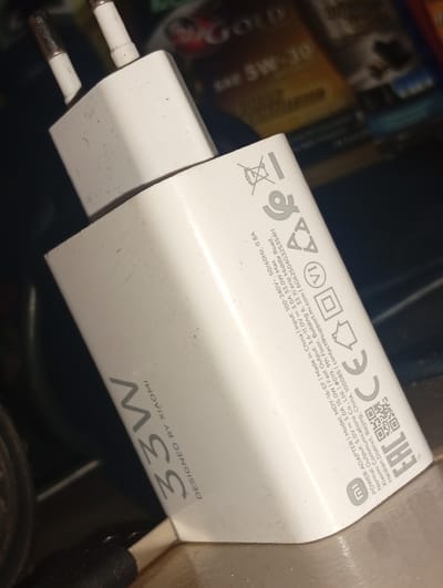 A white Xiaomi 33W fast charger is shown in the image. The charger has visible dust and smudges on its surface. The text "33W DESIGNED BY XIAOMI" is printed on the charger in a gray font. Other markings on the charger include various logos and technical specifications such as "Input: 100-240V~ 50/60HZ 0.8A" and "Output: 5V 3.0A 15.0W; 9V 2.23A 20.07W; 12V 1.67A 20.04W". The charger is positioned at an angle, with its European-style prongs pointing upwards. The background is dark and out of focus, showing some blurry items that appear to be other electronic devices or packaging. There is no indication of the location being Ukukwa, Nigeria.