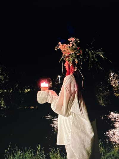 A person stands in profile on the grassy bank of a body of water at night. They are wearing a large floral headdress and a loose-fitting white garment. In their right hand, they hold a lit candle, the flame casting a warm glow. The background is dark, with the water reflecting some light, and trees or foliage are visible on the far bank. The overall scene suggests a ritualistic or ceremonial act. The specific location is within the municipality of Chotěšov, Czechia.