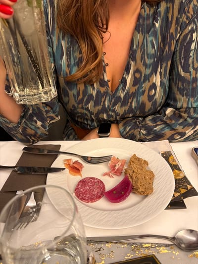 The image showcases a woman at a dinner table. She wears a patterned dress and a watch on her wrist. A plate of food rests beside her, containing bread, ham, beetroot, and salami. Silverware is arranged beside the plate. A large glass sits near her hand, with red nail polish visible on her fingers. The table is covered with a white tablecloth adorned with gold accents.