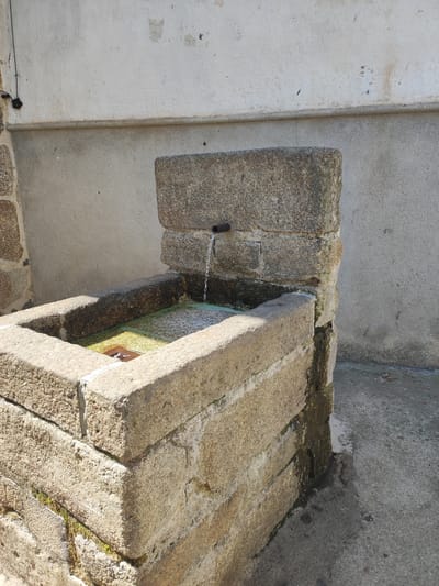 The image shows a stone fountain in Cadalso, Spain. Water is flowing from a pipe into a stone basin filled with green water. The fountain is constructed from rough, weathered stone blocks, with moss and algae visible on the surfaces. A concrete ground surrounds the fountain, and a weathered wall forms the background. The scene is illuminated by natural sunlight, casting shadows and highlighting the textures of the stone and concrete.