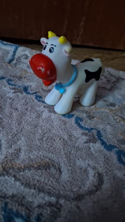 A toy cow is positioned on a textured rug. The toy is white with black spots on its back, yellow horns, a red nose, and a blue collar with a small blue bow. The rug has a light brown and gray base with thin, wavy blue lines. In the background, a dark wooden surface is visible. The scene appears to be indoors.