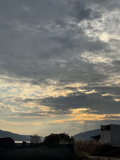 The image shows a cloudy sky over a coastal landscape in Mrčevac, Montenegro. In the distance, a plane is visible against the sky. The foreground features a fence, dry vegetation, and a partially visible building. Further back, a trailer and a red car are parked near the water, with hills forming the background. The lighting suggests it is either sunrise or sunset.