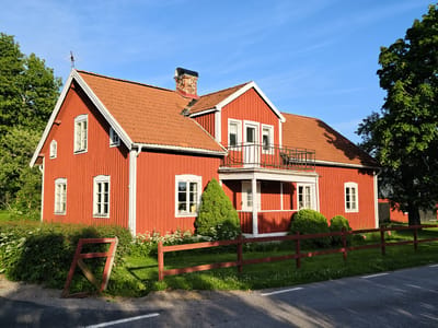A bright red wooden house with a terra cotta tile roof is the central subject in this media file, set against a clear blue sky. The house features white trim around its windows and doors, and a small balcony with a black railing overlooks a porch area. A chimney with a black cap rises from the roof.

The house is surrounded by lush greenery, including mature trees, bushes, and a flowerbed with white blossoms. A rustic, weathered wooden fence runs along the front of the property, separating it from a paved road. The fence is a reddish-brown color, with some sections appearing a bit dilapidated.

The scene suggests a peaceful rural setting. The sunlight casts distinct shadows, indicating that it is likely late afternoon or early morning. The overall impression is one of a well-maintained, idyllic home in a natural environment. No people are visible in the image, and there is no visible text. The location is Slaka, Sweden.