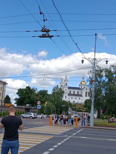 This is a medium shot of a street scene in Vitsebsk, Belarus, on a partly cloudy day. The foreground features a man in a black t-shirt and blue jeans standing at a crosswalk, looking away from the camera. Behind him, several people are walking across the street, some in groups. A white car and a silver SUV are visible on the road.
In the mid-ground, a white building with gold domes and spires, likely a church or cathedral, dominates the background. It is surrounded by lush green trees and a decorative lamppost with two lights. Overhead, power lines and their complex support structures are visible against the blue sky dotted with white clouds.
The activity captured is pedestrian movement across a busy intersection, with a mix of individuals and small groups navigating the crosswalk. The overall atmosphere appears to be one of everyday urban life, with a clear view of notable Belarusian architecture. The presence of power lines and the style of the lamppost suggest an urban or suburban setting. The weather appears pleasant, with a mix of sun and clouds. No text is clearly discernible in the image.