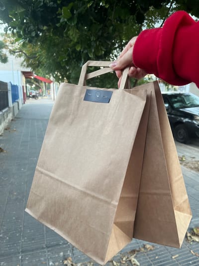 A person wearing a red sweater holds two brown paper bags with handles. One bag has a dark blue rectangular sticker with white text/graphic, possibly a logo. The background shows an urban street with trees, buildings, and parked cars. The location is San Fernando del Valle de Catamarca, Argentina. The ground appears to be a tiled sidewalk or walkway with debris.