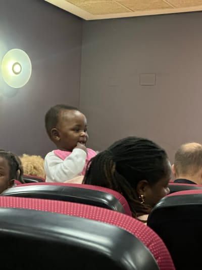 The image shows a baby with dark skin and short, dark hair, wearing a white and pink shirt. The baby is sitting in a seat on a train or bus, as suggested by the presence of other people, seatbacks with red patterned upholstery and black frames, and a round light fixture on the ceiling. A woman with dark, braided hair and gold earrings is sitting in the seat in front of the baby. Another person with short, gray hair is partially visible above the woman. The picture seems to be taken on public transportation, possibly a train or a bus. There is no information to confirm this is in Sant Vicent del Raspeig, Spain.