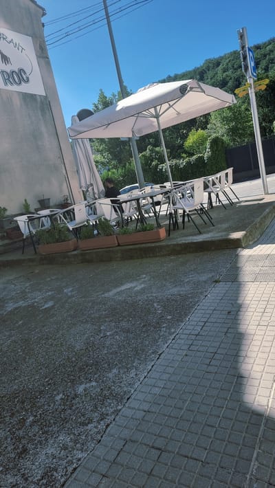 The image depicts the outdoor seating area of a restaurant. A large white umbrella shades several white tables and chairs. Planters with greenery are placed along the edge of the seating area. In the background, a wall with a sign reading "Restaurant Roc" is visible. There are a few people present, including one man sitting at a table. A street sign with directional arrows and a parking symbol is also visible on a pole to the right of the image. The scene appears to be set in a town with trees and a hillside in the background. The paving in the foreground is a mix of asphalt and square tiles. The sky is clear and blue. The overall impression is of a typical outdoor dining space on a sunny day.