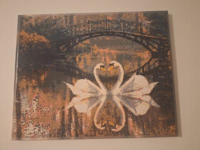 The image is a diamond painting of two swans with their necks intertwined, forming a heart shape. They are positioned in front of a dimly lit, possibly nighttime, scene with a bridge arching over water. The background appears to be a forest or park with warm, muted tones of brown, orange, and dark blue. The overall impression is romantic and serene. The diamond painting is framed with a metallic border and hangs on a plain, light-colored wall. There are no people or animals present in the scene other than the depicted swans. The image is a static depiction, and no actions or interactions are occurring. There is no information available to place this artwork in the city of Mińsk Mazowiecki, Poland.