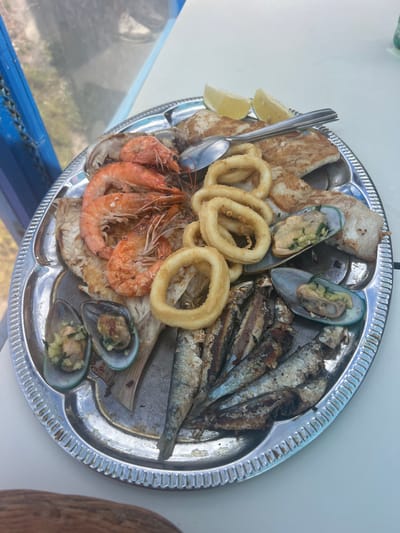 A silver platter is filled with a variety of seafood, including several large cooked shrimp, fried calamari rings, whole grilled sardines, mussels in their shells, and pieces of grilled fish. The seafood is arranged on the platter, with lemon wedges and cutlery placed amongst the food. The platter sits on a white table, with a blurred background suggesting an outdoor setting with some blue structures and greenery. The lighting indicates it is daytime.