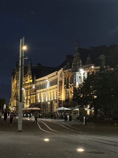 The image captures a nighttime view of a city street in Ghent, Belgium. In the foreground, a paved street with tram tracks is visible, illuminated by embedded lights in the pavement and a tall streetlamp to the left. Several people are walking or standing on the sidewalk. Behind them, a row of historic buildings with ornate facades and illuminated windows line the street. Some of these buildings appear to be restaurants or cafes with outdoor seating areas covered by umbrellas. The sky above is dark, indicating it is nighttime. The overall scene conveys a lively urban atmosphere at night.