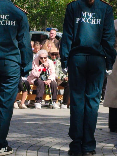 The image captures a moment outdoors, likely in a public space in Apatity, Russia, given the presence of Cyrillic lettering on the uniforms. In the foreground, two individuals in dark blue uniforms with white Cyrillic lettering that reads "МЧС РОССИИ" (MChS ROSSII), which translates to "Ministry of Emergency Situations of Russia," are standing. Their backs are to the camera, with the person on the right prominently displaying the lettering.

In the middle ground, a group of people are seated on a wooden bench. An elderly woman in a light pink jacket and dark pants is in the center, holding a cane and a bouquet of red flowers. She is wearing sunglasses. To her left, another elderly woman is also seated, wearing a brown hat and a green scarf. Several other individuals, including children and adults, are visible behind them. A white van with a blue light bar on top is parked behind the seated group, suggesting a possible event or official gathering.

The ground is paved with interlocking bricks, and in the background, trees and foliage are visible, indicating an outdoor setting in what appears to be a park or a town square. The lighting suggests it is daytime, with sunlight casting shadows on the ground. The overall scene suggests a civic event or commemoration, possibly involving the Ministry of Emergency Situations.